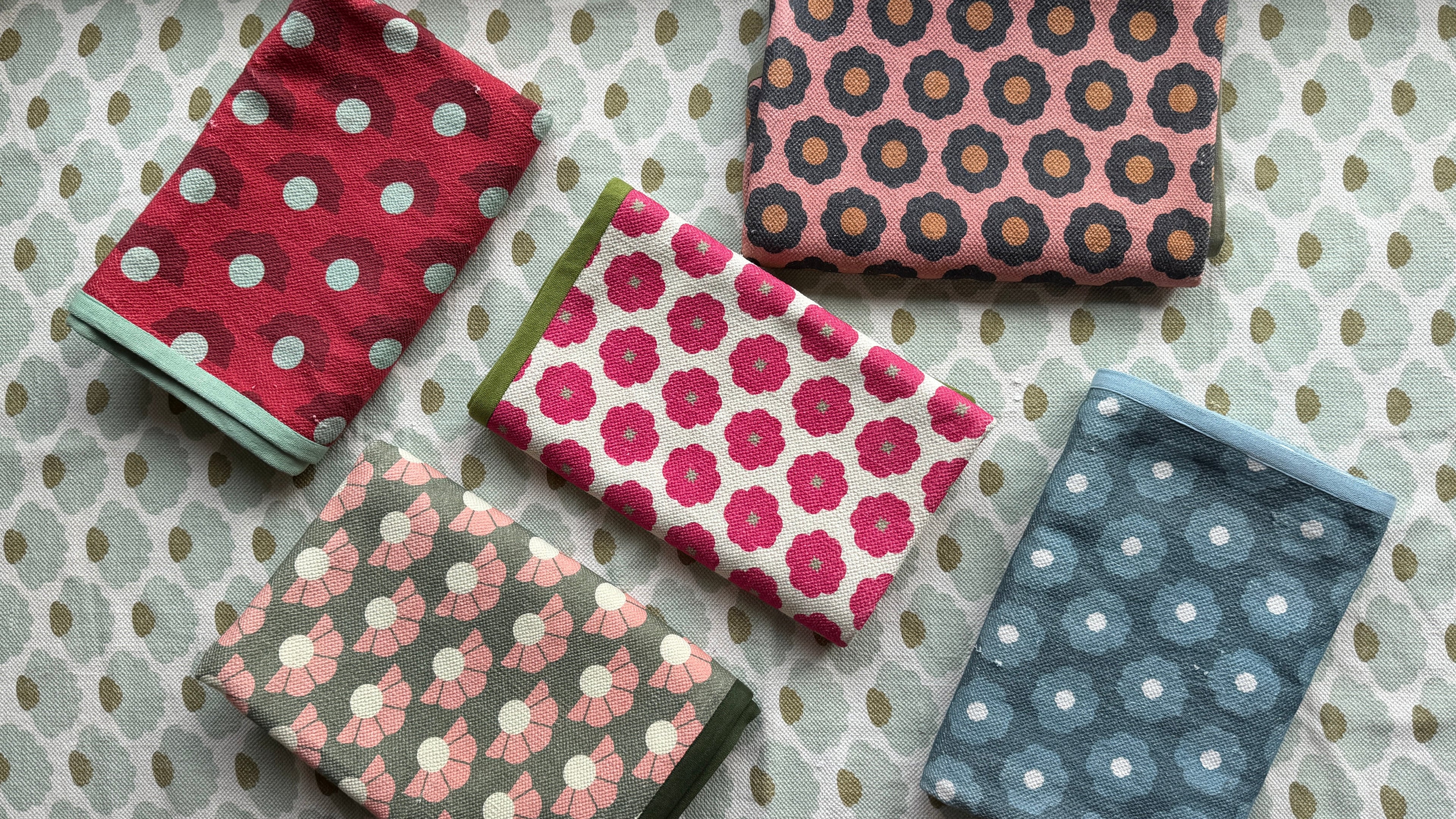 Four folded cotton rugs with different floral and polka dot patterns in pink, blue, red, and green, arranged on a patterned background.